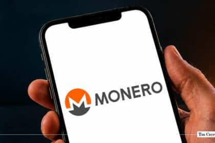 Hand holding a smartphone displaying the Monero logo and wordmark on a bright white screen