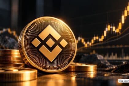 Physical Binance Coin (BNB) token displayed on a dark surface in front of a golden candlestick trading chart.