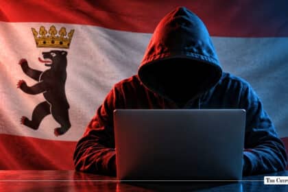 Individual in a dark hooded sweatshirt sitting in front of a laptop computer, silhouetted against an illuminated German state flag of Berlin