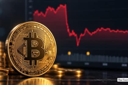 Gold Bitcoin token standing upright next to a stack of coins against a digital screen displaying a sharp red downward price chart