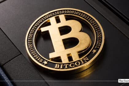 Gold-plated Bitcoin coin featuring binary code engravings on its outer ring resting on a dark, textured surface
