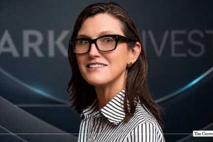 Cathie Wood, CEO and CIO of ARK Invest, wearing black glasses and a striped shirt against an ARK Invest branded background
