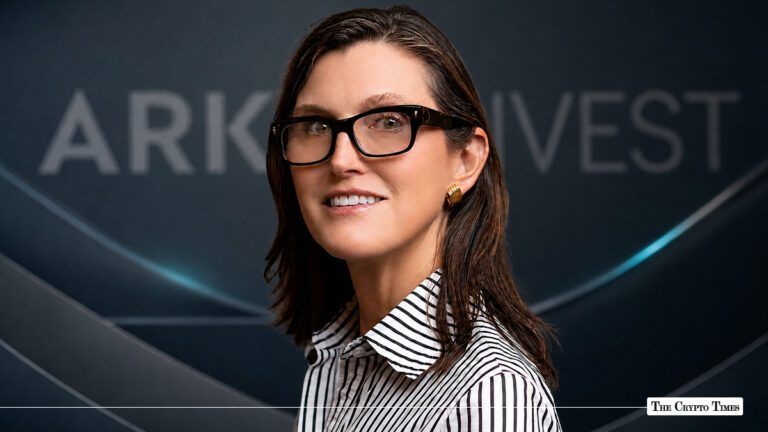 Cathie Wood, CEO and CIO of ARK Invest, wearing black glasses and a striped shirt against an ARK Invest branded background