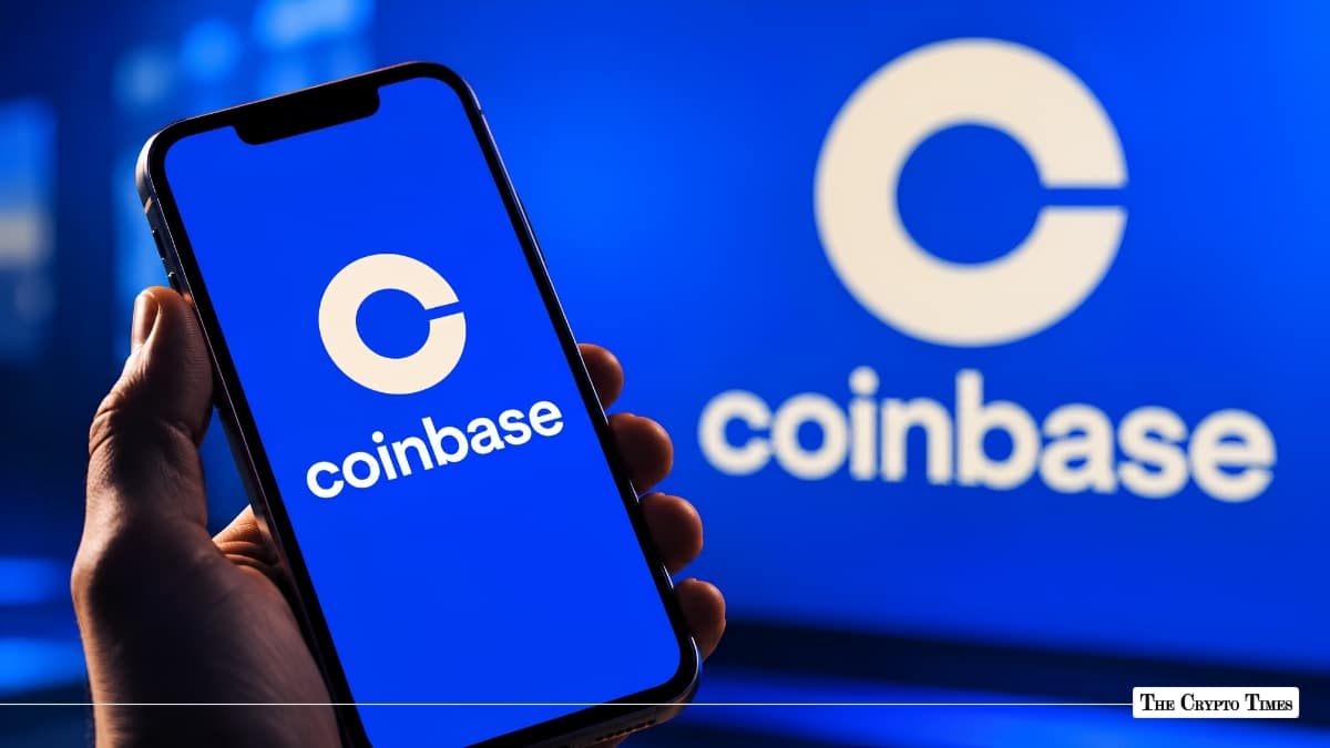 Hand holding a smartphone displaying the Coinbase app in front of a blue Coinbase screen background.