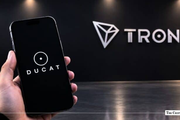 Hand holding a smartphone displaying the DUCAT logo in front of a Tron wall sign.
