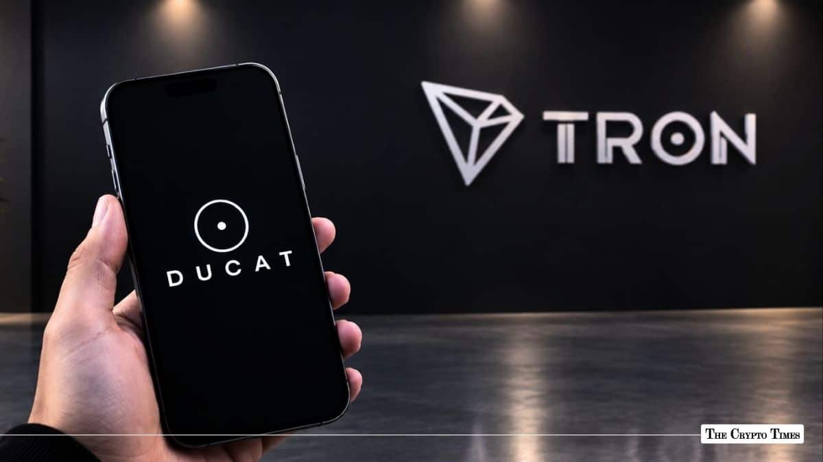 Hand holding a smartphone displaying the DUCAT logo in front of a Tron wall sign.