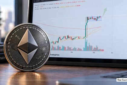 Physical silver Ethereum coin sitting on a wooden desk next to a laptop displaying an upward-trending candlestick chart.