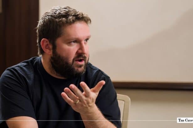 Man with a beard in a black t-shirt speaking with hand gestures during an interview.