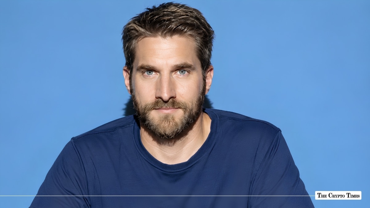 Studio headshot of a man with short brown hair and a beard against a blue background.