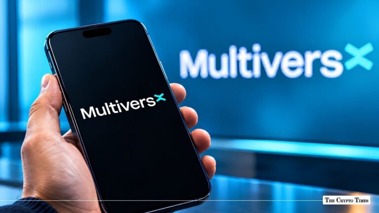 Hand holding a smartphone displaying the MultiversX logo in front of a blue brand monitor.