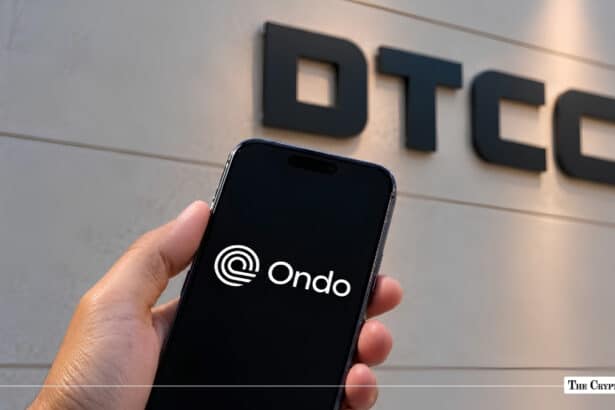 Hand holding smartphone displaying the Ondo Finance logo in front of DTCC wall signage.