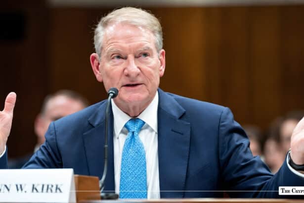 Official speaking into microphone with open hands at a hearing desk behind a nameplate reading "N W. KIRK".