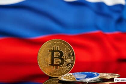 A gold Bitcoin coin standing upright next to smaller crypto coins, set against the blurred red, white, and blue colors of the Russian national flag