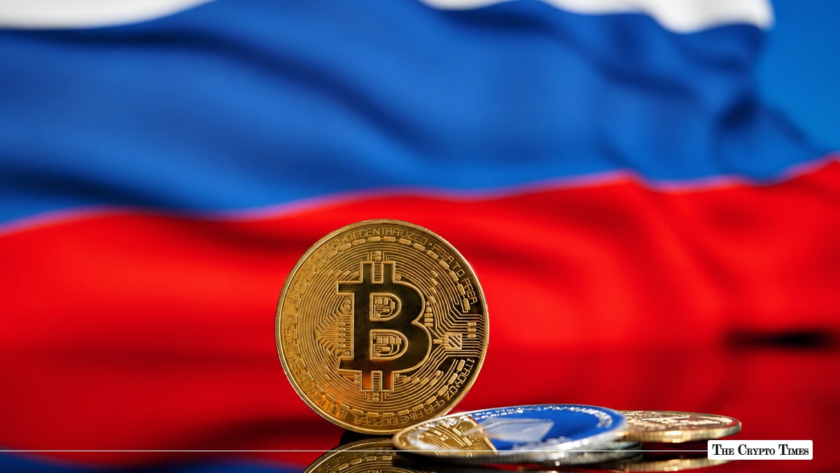 A gold Bitcoin coin standing upright next to smaller crypto coins, set against the blurred red, white, and blue colors of the Russian national flag