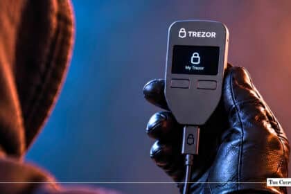 Gloved hand holding a illuminated Trezor hardware wallet displaying "My Trezor" next to a hooded figure