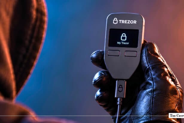 Gloved hand holding a illuminated Trezor hardware wallet displaying "My Trezor" next to a hooded figure