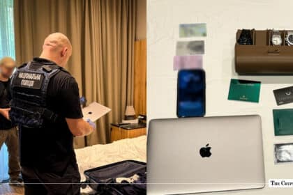 Ukrainian police officer inspecting a laptop during a room raid next to seized evidence including luxury watches, bank cards, and electronics.