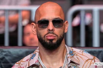 Andrew Tate wearing sunglasses and silver chains sitting ringside at a public event
