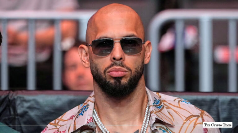 Andrew Tate wearing sunglasses and silver chains sitting ringside at a public event