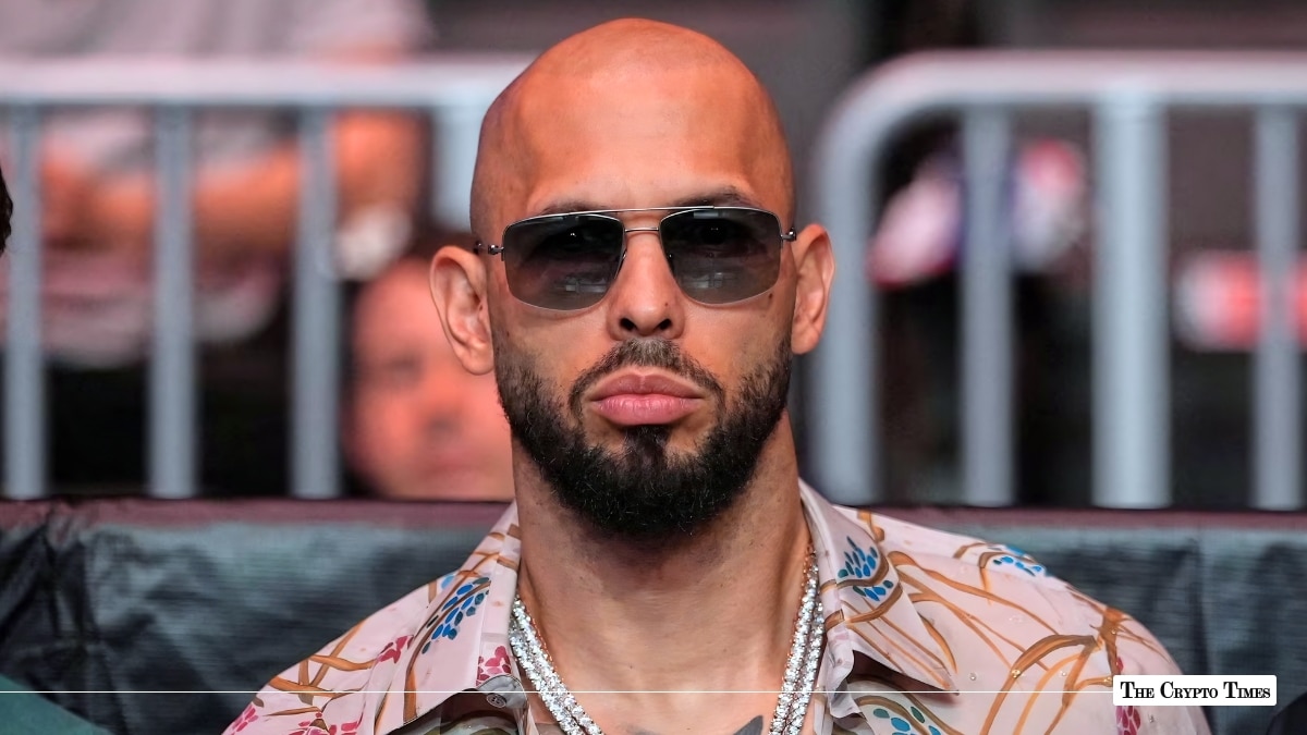 Andrew Tate wearing sunglasses and silver chains sitting ringside at a public event
