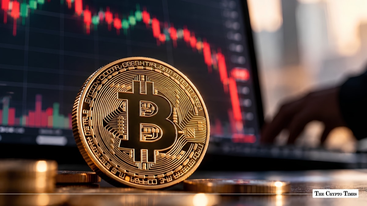 Physical Bitcoin coin resting in front of a laptop displaying a downward-trending red candlestick chart.