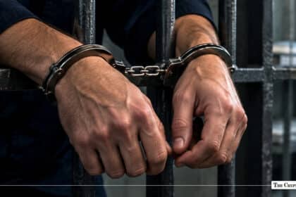 Close-up of a person's handcuffed hands resting against the dark metal bars of a jail cell