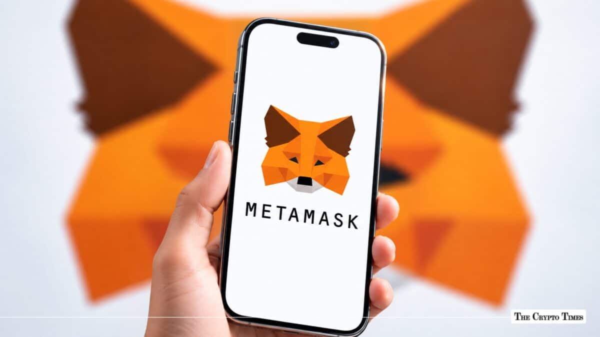 A hand holding a smartphone displaying the MetaMask logo in front of a blurred MetaMask fox wall art background.