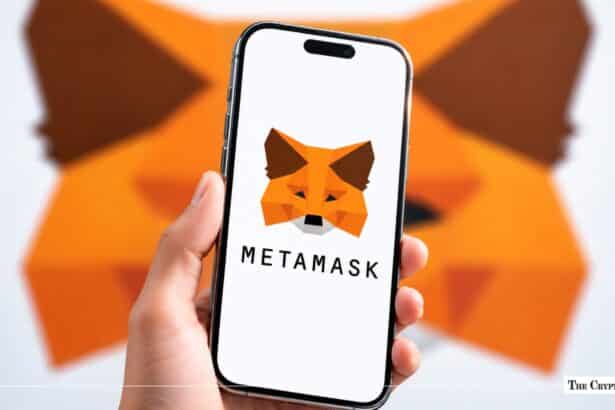 A hand holding a smartphone displaying the MetaMask logo in front of a blurred MetaMask fox wall art background.