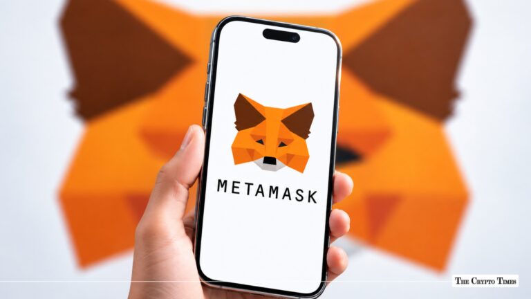 A hand holding a smartphone displaying the MetaMask logo in front of a blurred MetaMask fox wall art background.