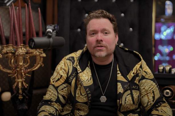 Richard Heart speaking in front of a microphone wearing a gold patterned Versace hoodie and HEX pendant.