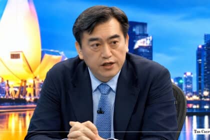 Man in a dark suit speaking in a studio setting with the Singapore skyline background.