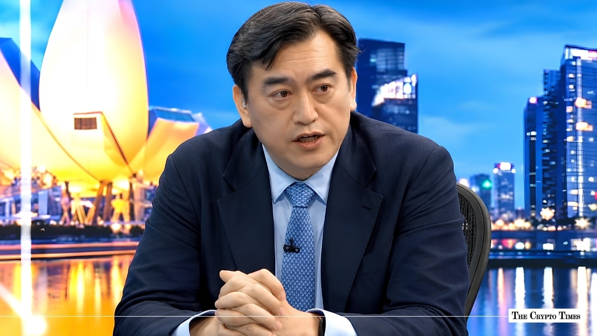 Man in a dark suit speaking in a studio setting with the Singapore skyline background.