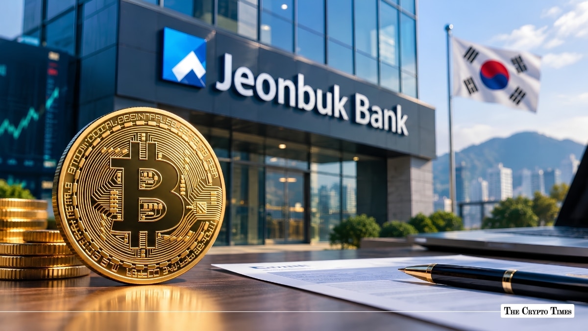 Physical Bitcoin coin next to documents in front of Jeonbuk Bank building and South Korean flag.