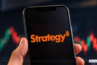Hand holding a smartphone displaying the orange MicroStrategy logo with a Bitcoin symbol superscript against a stock chart background