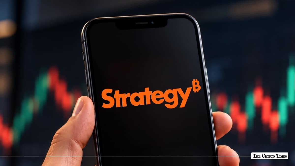 Hand holding a smartphone displaying the orange MicroStrategy logo with a Bitcoin symbol superscript against a stock chart background