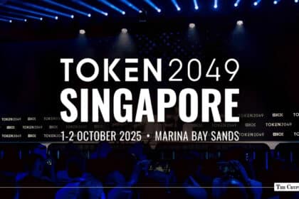 TOKEN2049 Singapore event backdrop on a dark stage displaying "1-2 October 2025 • Marina Bay Sands" with blue spotlights and audience silhouettes below