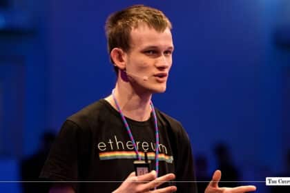 Ethereum co-founder Vitalik Buterin gesturing while speaking on stage in a black Ethereum t-shirt against a dark blue background