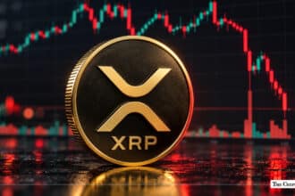 Physical gold XRP coin standing upright in front of a market candlestick chart.
